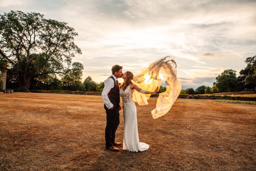 Bride and Groom kiss with veil billowing in the sunset at Braxted Park