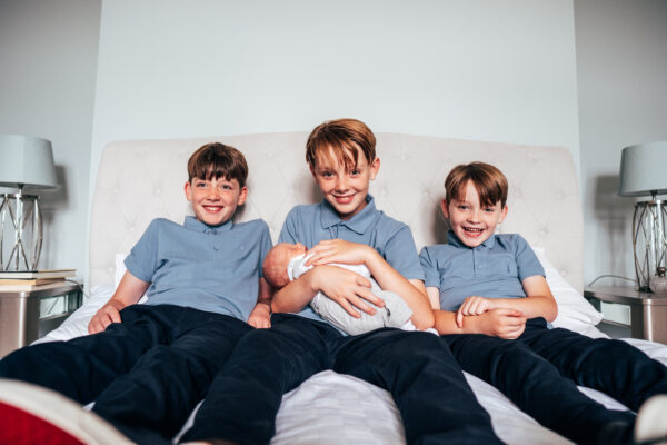 Three boys in matching outfits smile to camera as the eldest holds new baby brother Essex at home lifestyle family childhood photographer