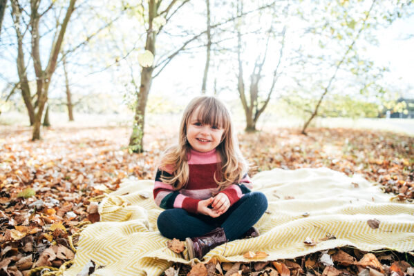 little girl in knitted dress sits on yellow blanket in autumn leaves Essex family childhood baby photographer