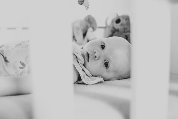 Baby girl looks at camera through bars of her cot at home lifestyle family newborn childhood photographer