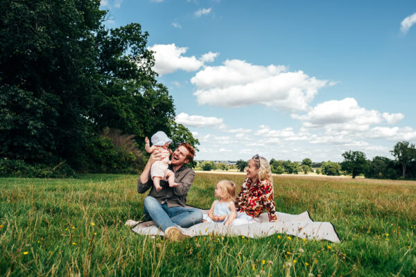 Family of four sit on blanket as dad lifts baby girl in the air at Hylands House Essex family lifestyle childhood baby photographer