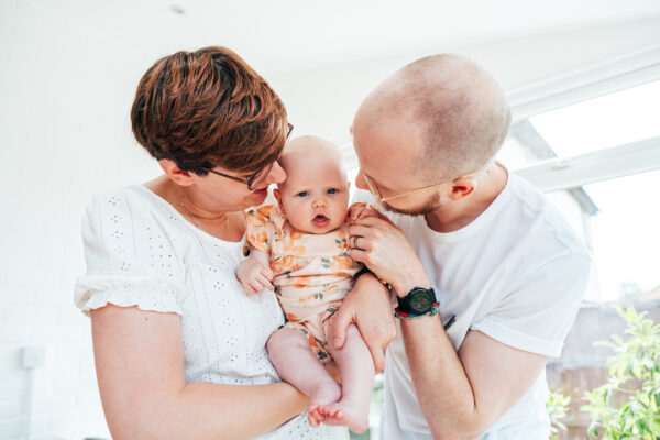 Mum and dad hold baby girl in orange between them in their conservatory at home Essex lifestyle childhood baby photographer