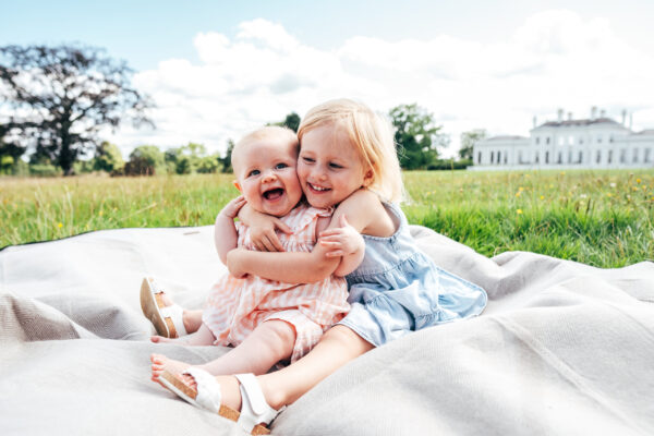 little girl cuddles smiling baby sister on the lawn outside Hylands House Essex lifestyle family baby childhood photographer