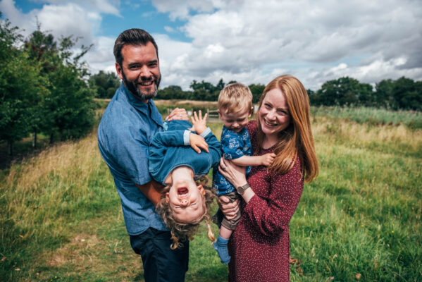 Family of four laugh in a field as dad holds daughter up side down Essex lifestyle family baby childhood photographer