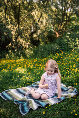 Little girls sits on blanket and picks buttercup in sunny field Essex family lifestyle childhood baby photographer