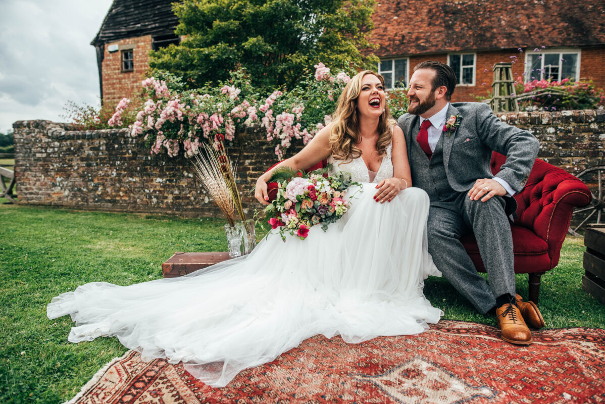 bride and groom sit on a red sofa and laugh at The House Meadow Best Essex Kent documentary wedding photographer