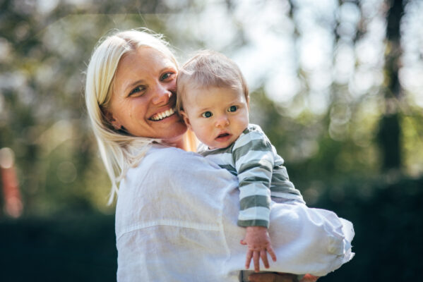 Mum holds up baby boy and smiles in sunny park Essex family lifestyle baby childhood photographer