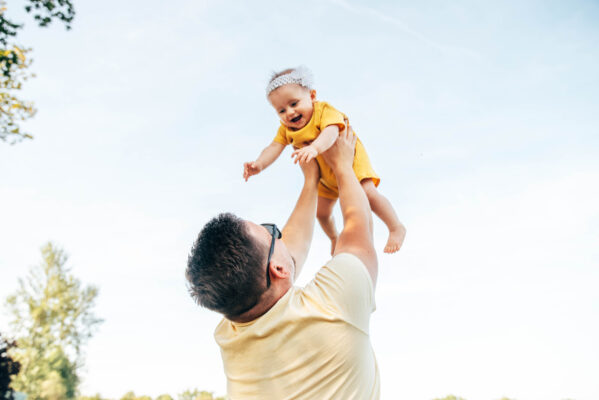 Dad lifts baby girl in yellow romper into the air as she laughs Essex lifestyle family childhood photographer