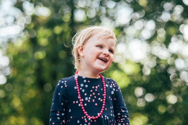 Little girl with pink necklace laughs in sunny park Essex family lifestyle childhood baby photographer