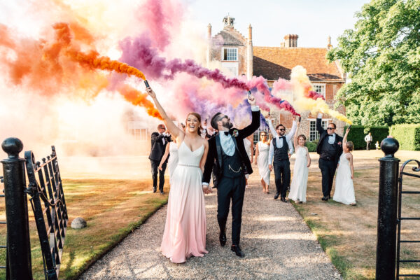 bride in pink and groom in black tie hold colourful smoke bombs with wedding party at Bruisyard Best Essex documentary wedding photographer