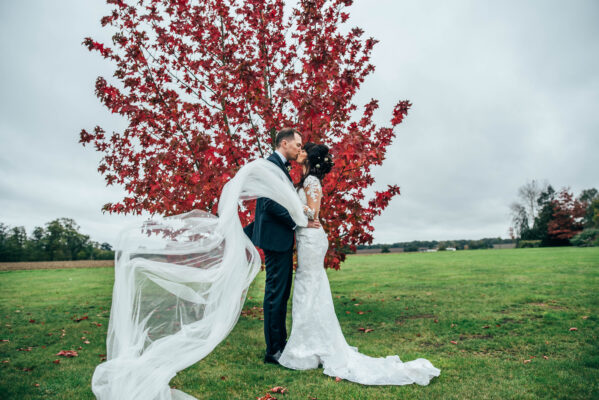 Bride and groom kiss in front of a red tree as her veil blows away in the wind at Blake Hall Best Essex documentary wedding photographer