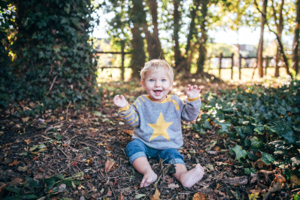 Little boy in knitted jumper sits amongst autumn leaves and smiles Essex lifestyle family baby childhood photographer