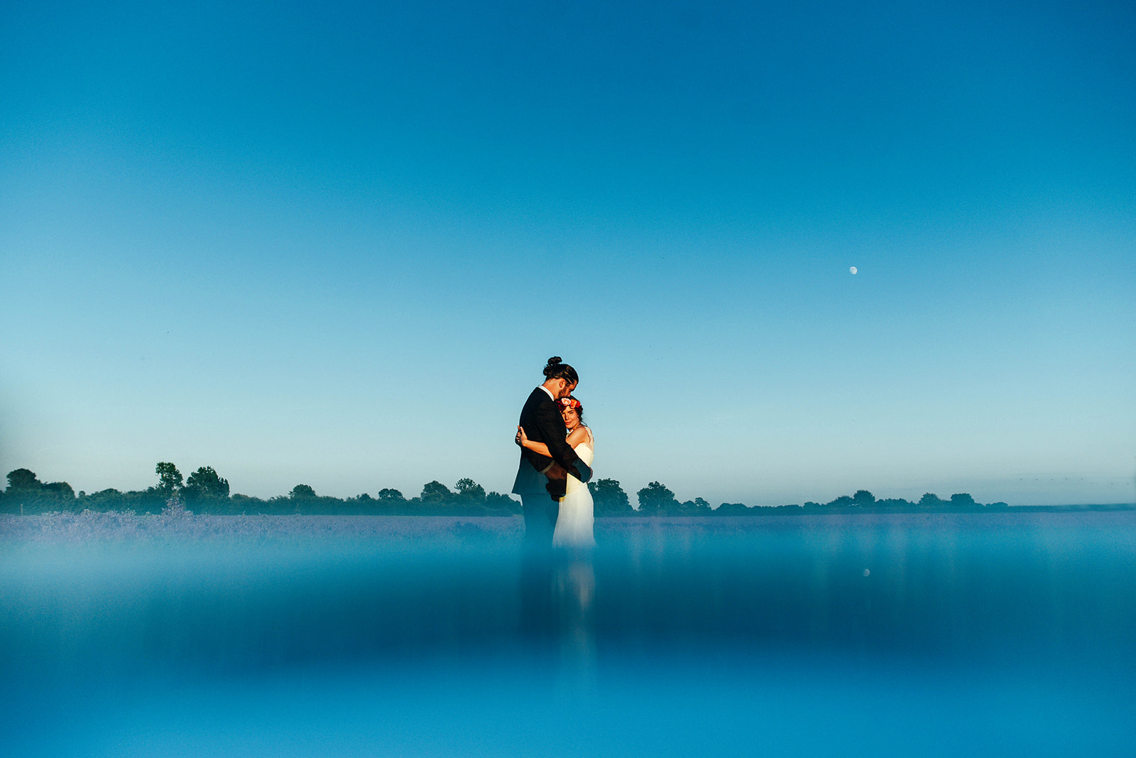 Groom in black tie hugs bride in flower crown in a field at blue hour. Houchins. Best Essex documentary wedding photographer