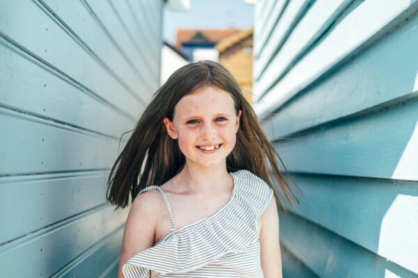 little girl smiles as hair blows in breeze in-between two blue beach huts Essex lifestyle family childhood baby photographer