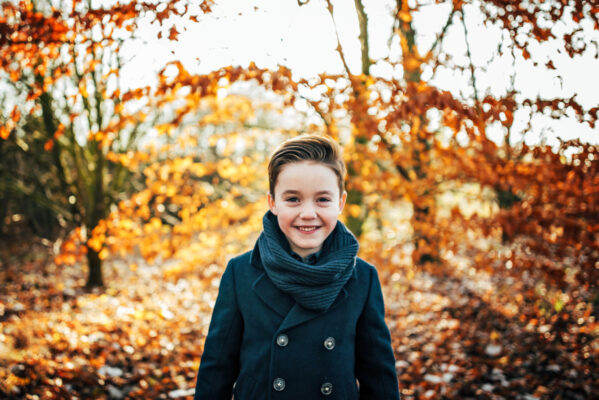 Boy in navy coat and scarf smiles in a sunlit field in autumn Essex family lifestyle childhood baby photographer