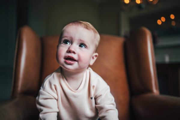 Baby girl sits on leather wingback chair at home Essex lifestyle family baby childhood photographer