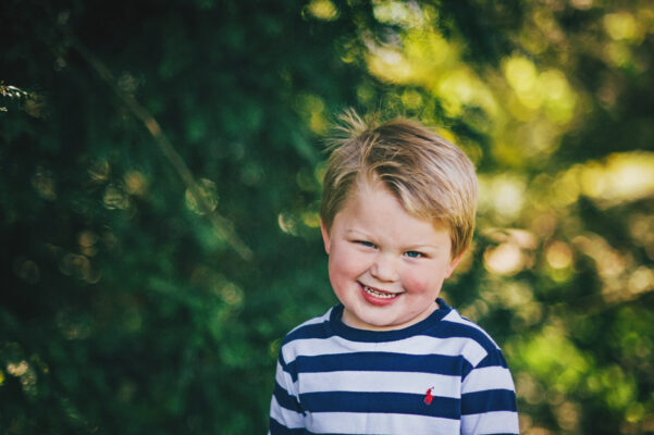 little boy grins at camera in sunny park Essex family lifestyle baby childhood photographer