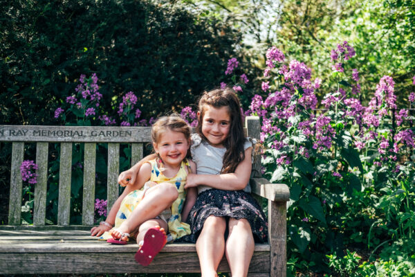 Two sisters hug on a bench in sunny park Essex family lifestyle childhood baby photographer