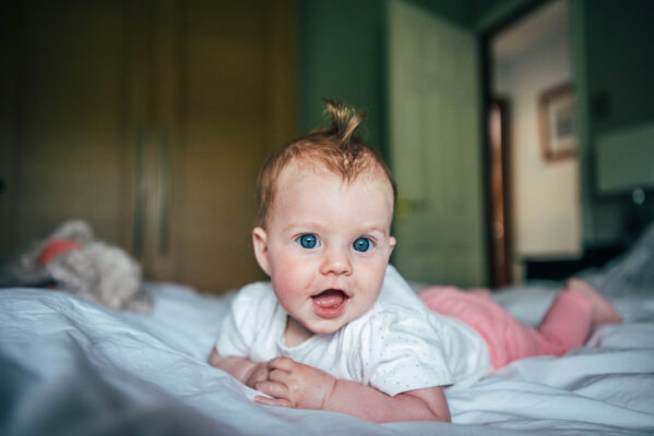 Little girl with tuft of red hair smiles as she lays on bed at home Essex family lifestyle baby childhood photographer