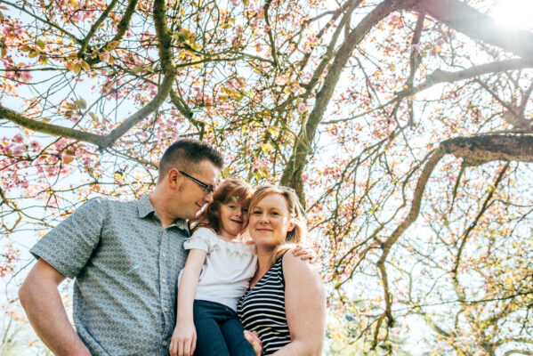 family of three cuddle under pink blossom tree on sunny day Essex family lifestyle baby childhood photographer