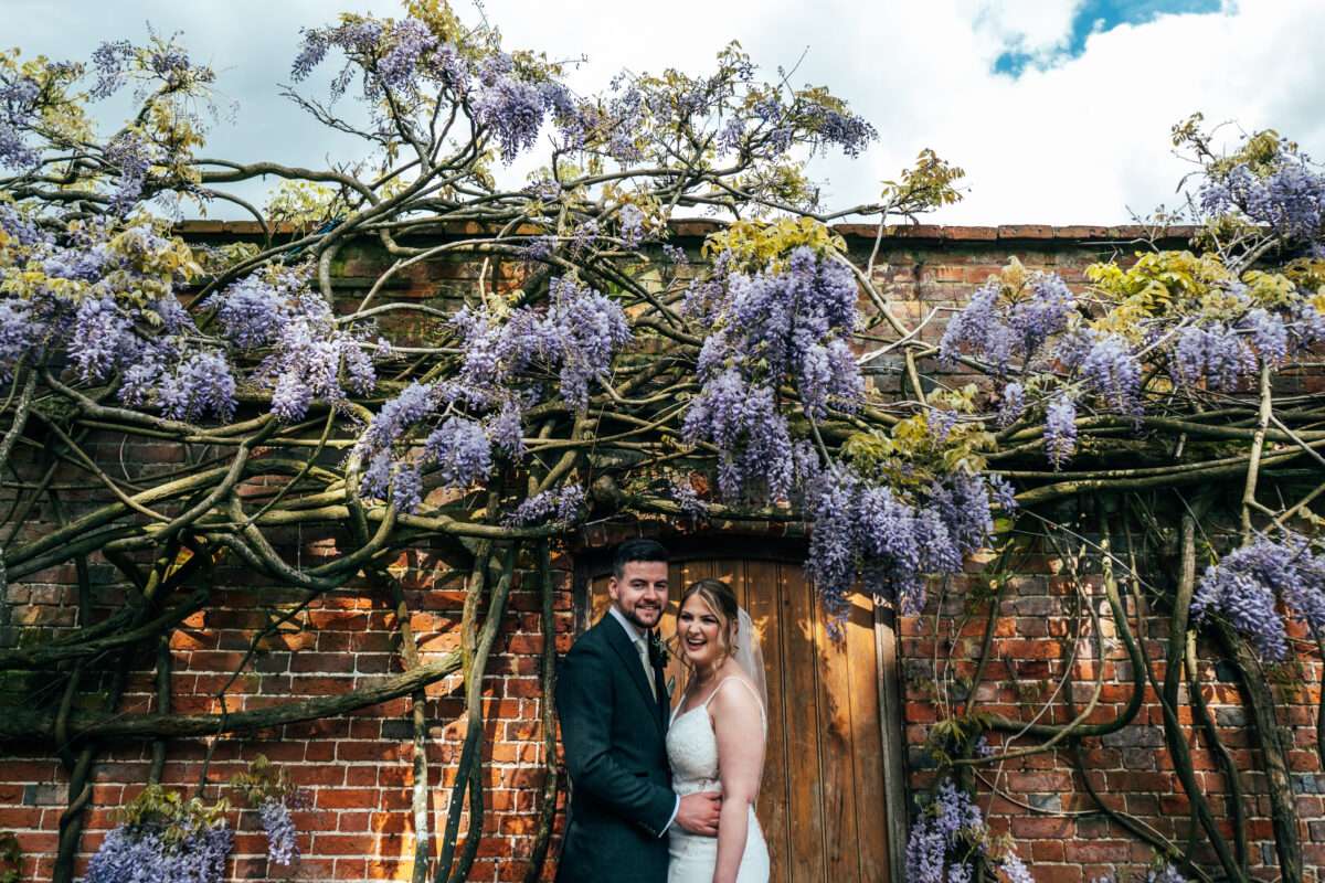 Bride & Groom stand under spring wisteria in walled garden at Blake Hall