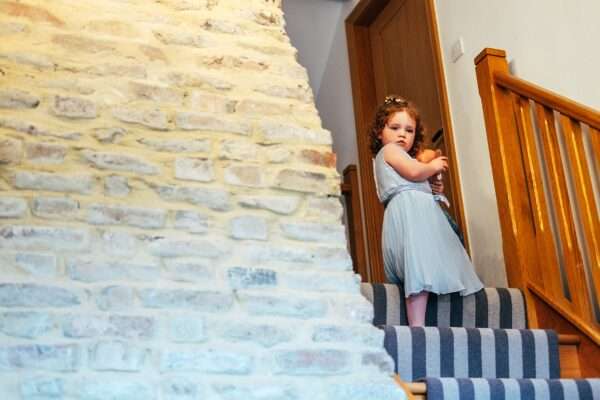 Flower girl in blue dress holds dolly at the top of stairs during prep for Captains Wood Barn wedding