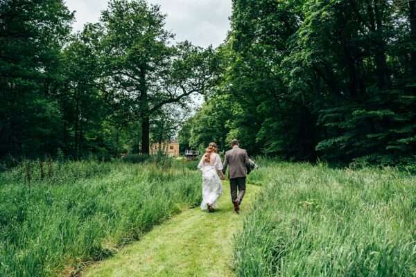 bride and groom walk through long grass and tall trees to drinks reception at Captains Wood Barn