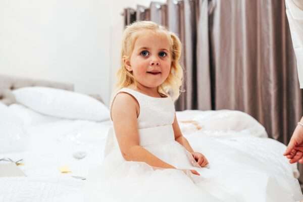 Flower girl sits on bed in white dress