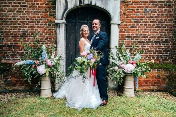 Bride and Groom smile in front of church door and floral display in Essex Church wedding