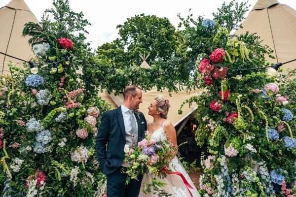 Bride and Groom under summer brights floral arch at tipi entrance for Essex back garden wedding