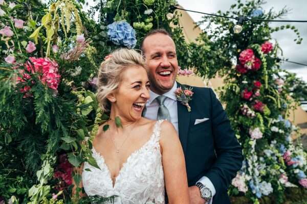 Bride and Groom laugh under big summer brights floral arch for back garden tipi wedding