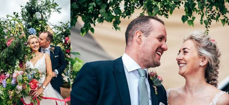 laughing bride and groom next to big summer bright floral arch for Essex back garden tipi wedding