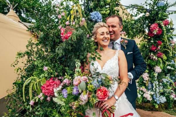 laughing bride and groom next to big summer brights floral arch for a back garden Essex tipi wedding