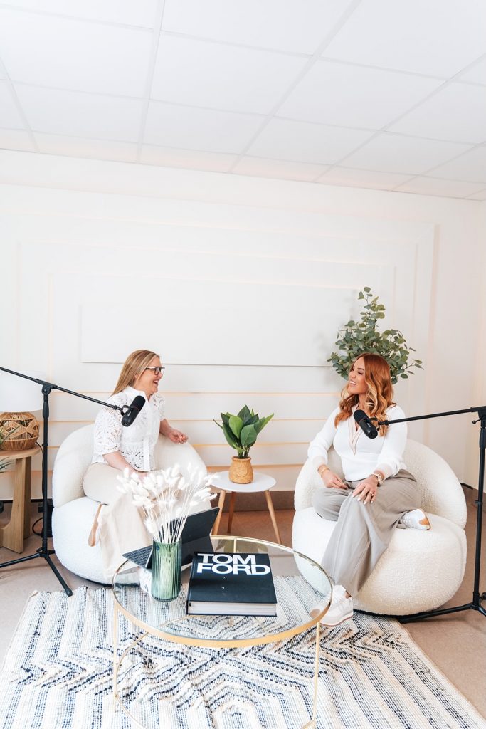 Two women in neutral setting talk into mics for podcast