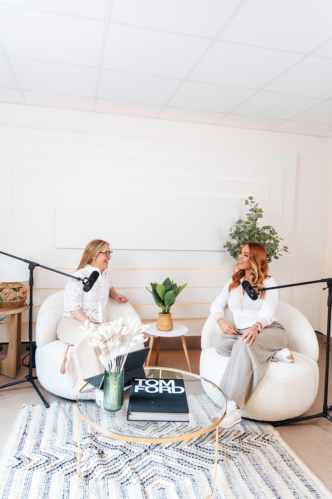 Two women in neutral setting talk into mics for podcast
