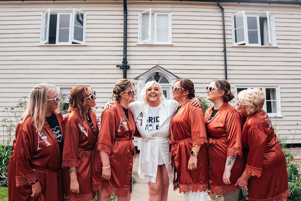 Smiling bride hugs bridesmaids in copper dressing gowns, all wear heart shaped sunglasses