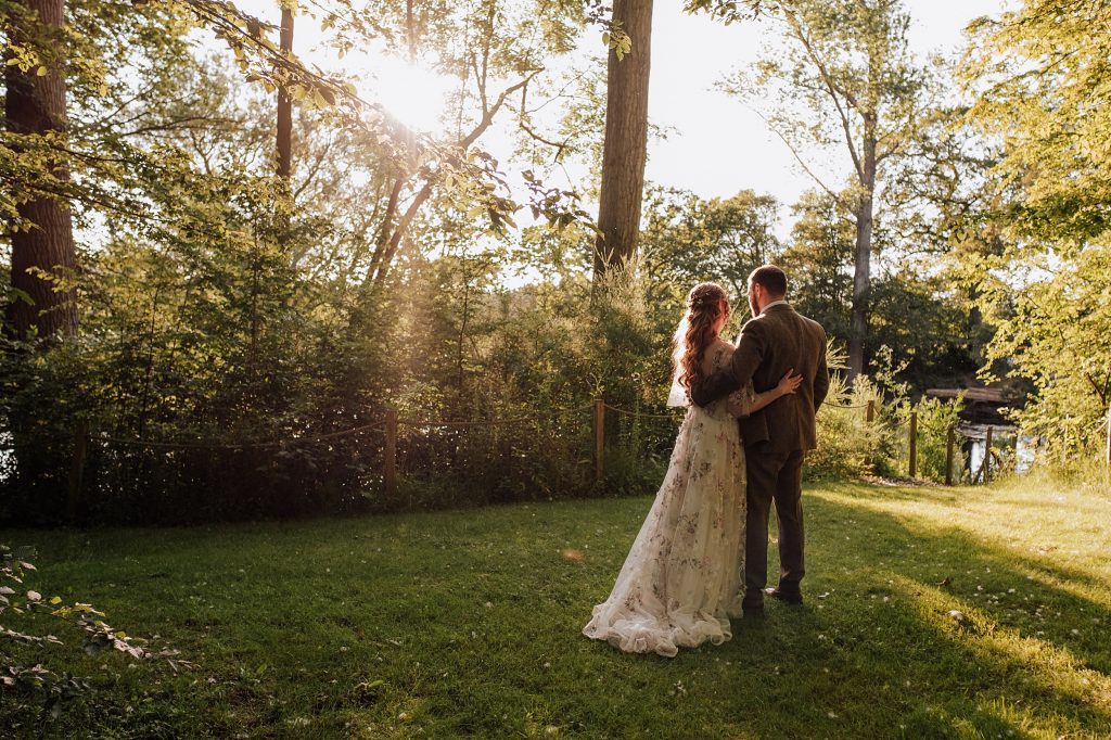 bride and groom stand facing away from camera as sunsets over treelike