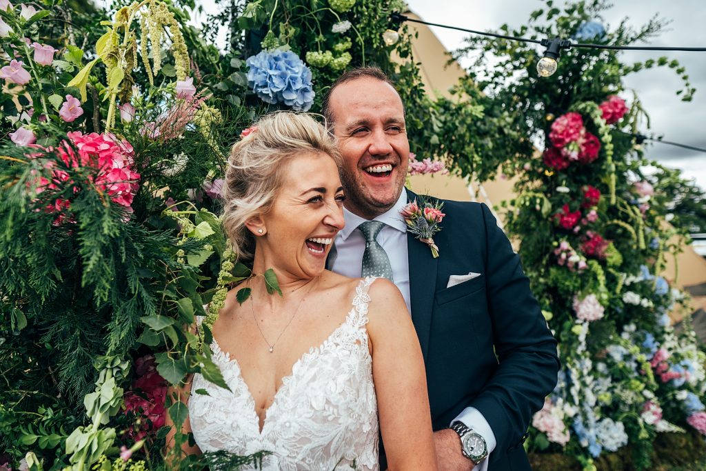 laughing bride and groom in front of floral arch