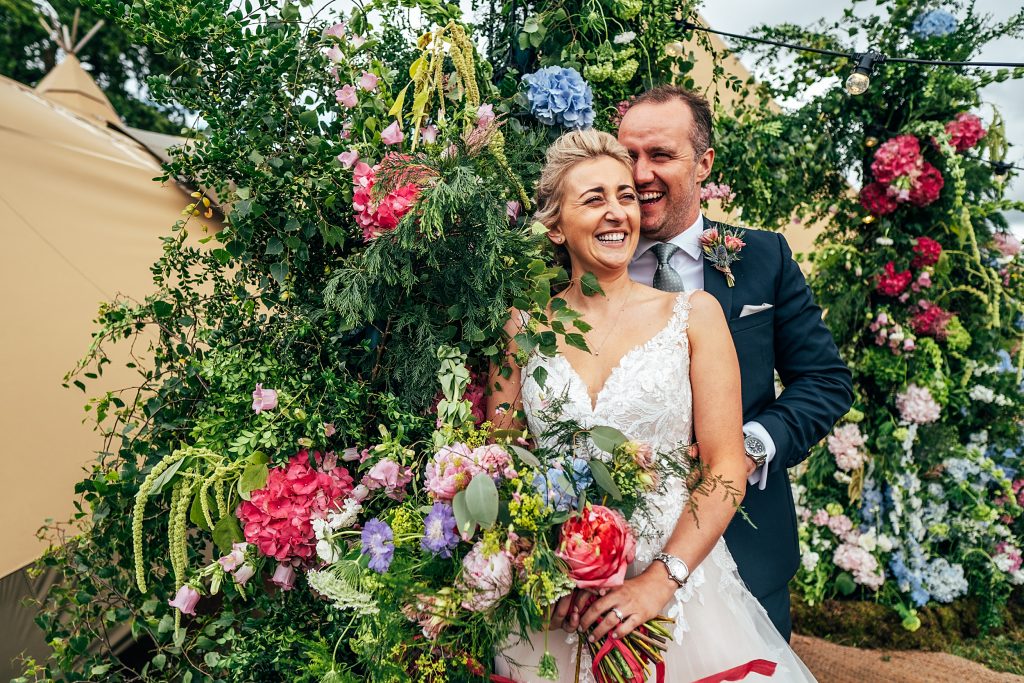 laughing bride and groom in front of floral arch outside tipi