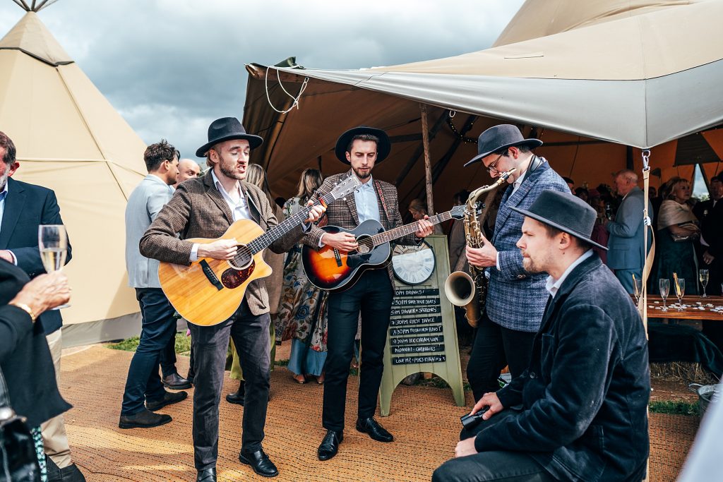 folk band in hats and tweed sing and play guitar outside tipi