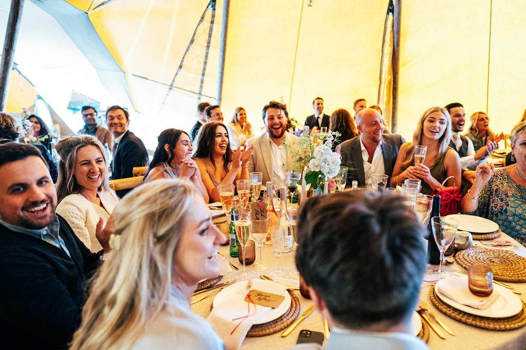 wedding guests round a table laugh at speeches in a tipi