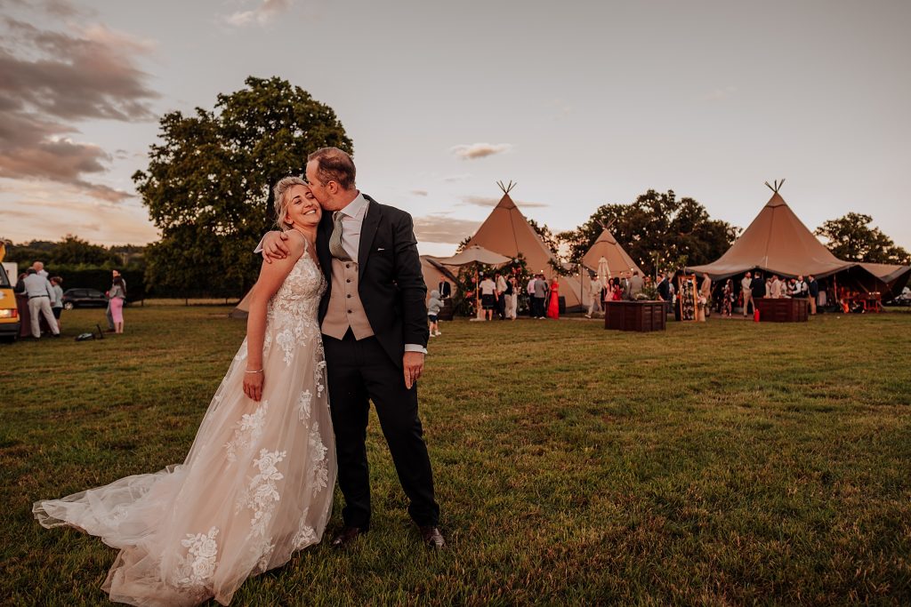 groom kisses bride in field at sunset behind them is tipi with wedding guests outside