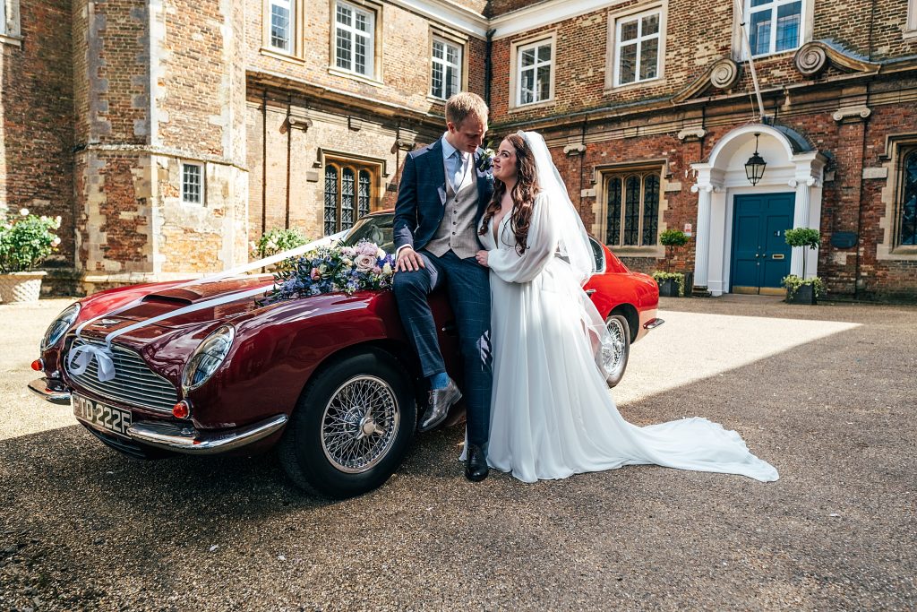 groom sits on bonnet of red sports car as bride leans in close