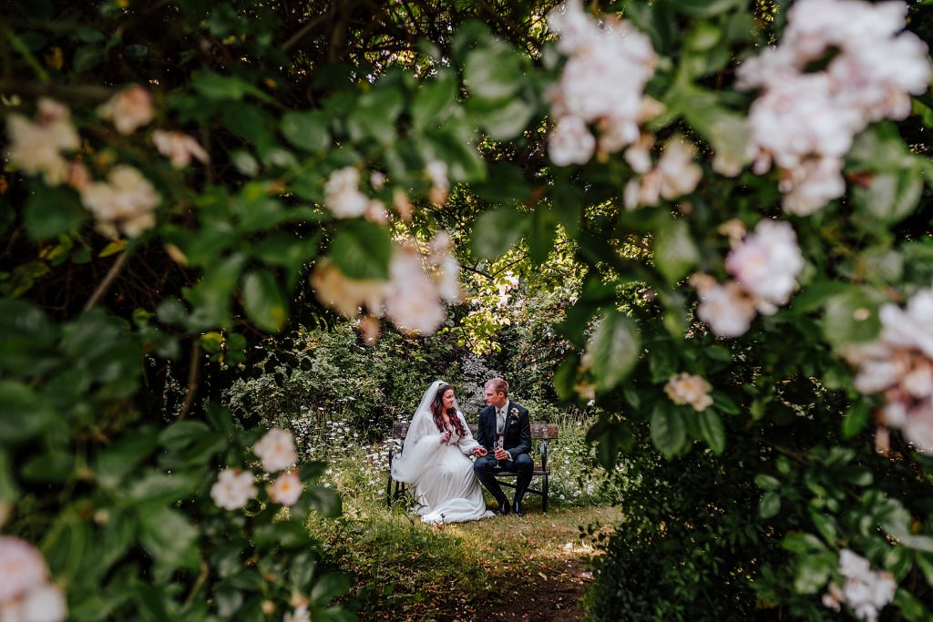 bride and groom sit on bench viewed through gap in blossom tree