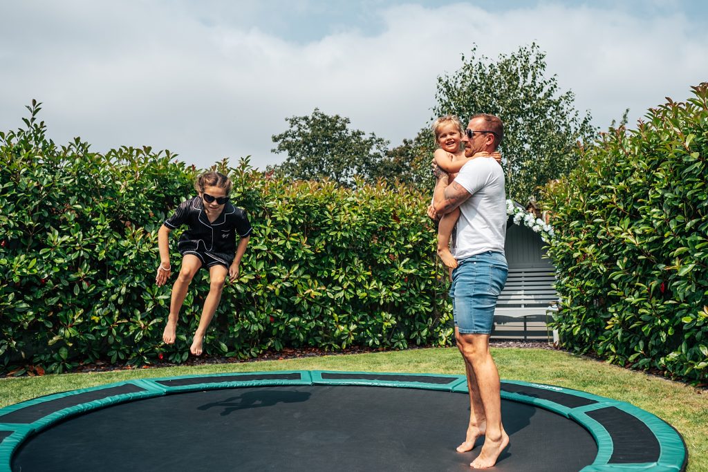 bridesmaid in black pyjamas and heart sunglasses jumps on a trampoline with man and baby in a garden