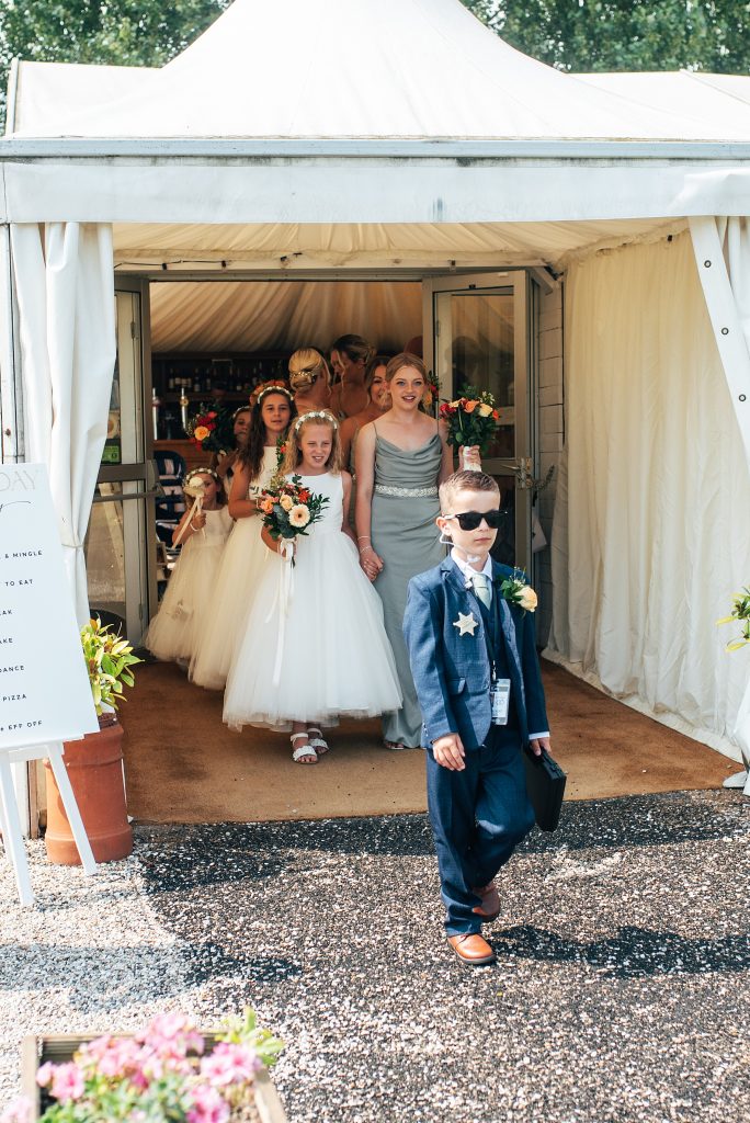 little boy in suit and glassed carries a briefcase leads bridesmaids and flower girls out of a marquee
