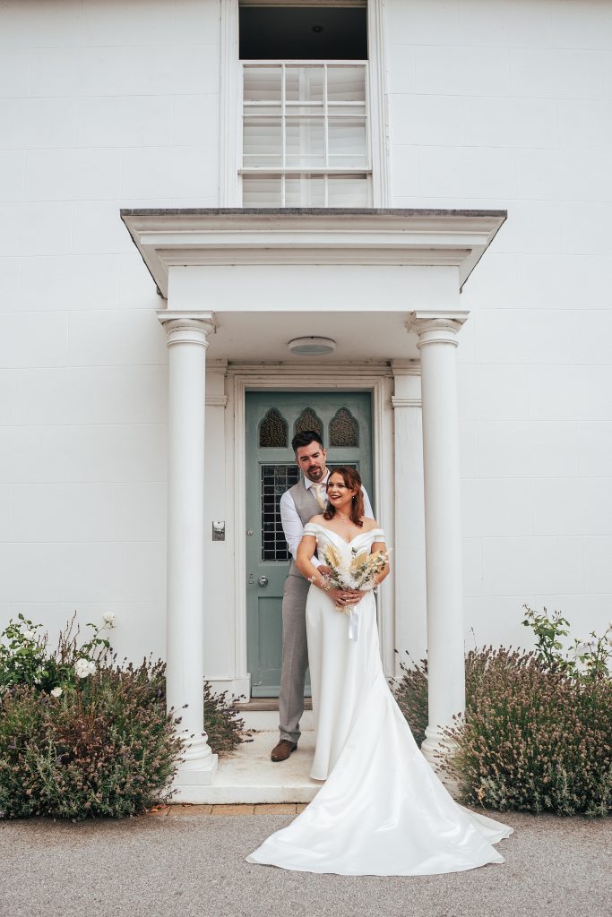 bride and groom stand on portico of big white house