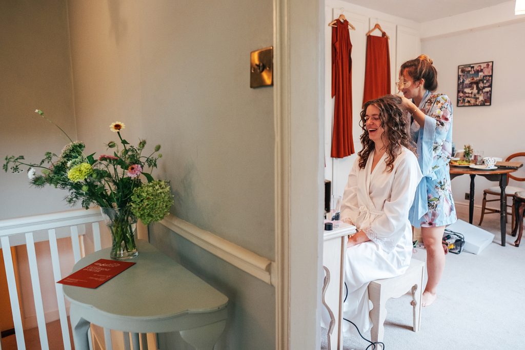 bride laughs as she has her hair done, bridesmaids dresses hang in the background