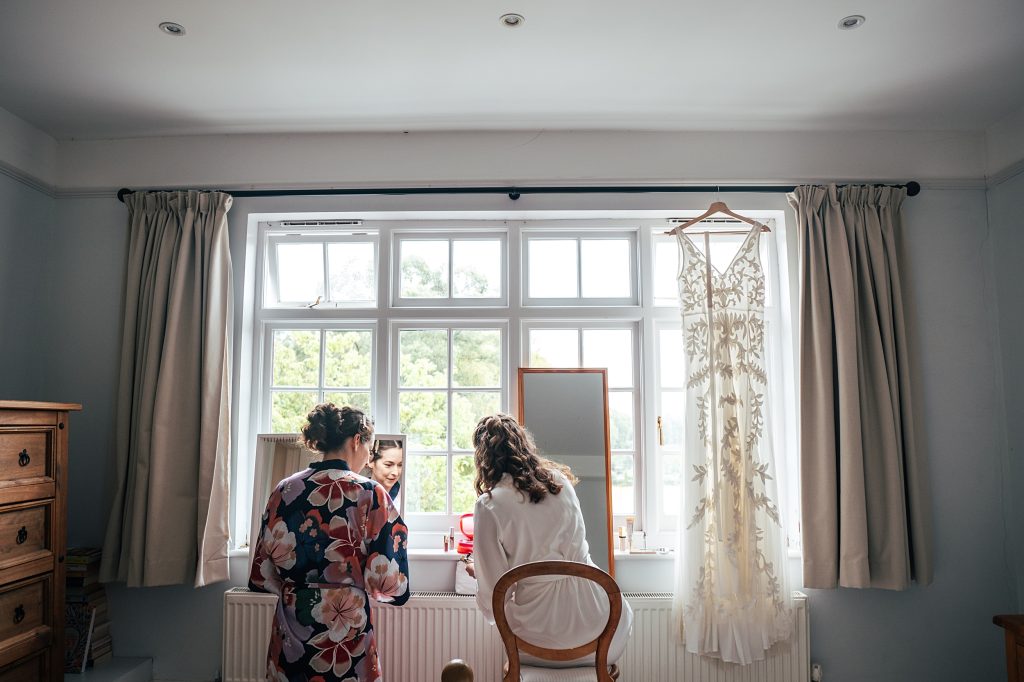 bride and bridesmaid do make up in front of a window, her dress hangs from curtain pole