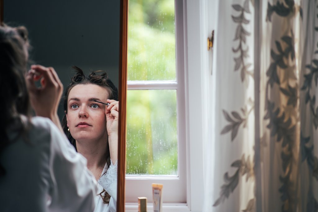 Bride does make up in mirror as rain falls outside the window